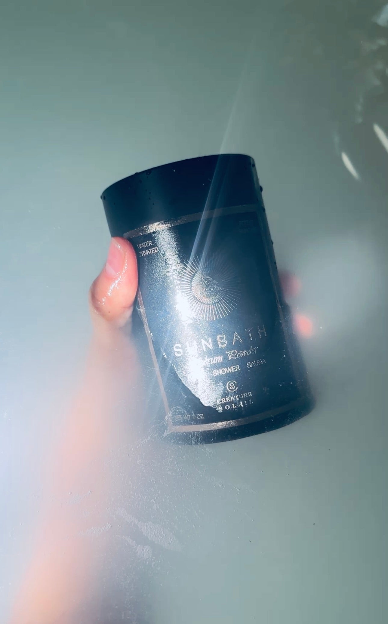 Hand holding a SUNBATH Serum Powder tin in a misty bath